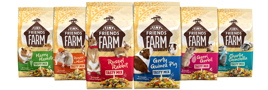 A horizontal lineup of six “Tiny Friends Farm” Tasty Mix pet food bags against a white background. From left to right: Green‑banded “Harry Hamster” mix with a golden hamster; Orange‑banded “Reggie & Mimi Rat” mix with two pet rats, one wearing a tiny cap; Red‑banded “Russel Rabbit” mix featuring an upright brown-and-white rabbit in a red bow tie; Blue‑banded “Gerty Guinea Pig” mix with a brown-and-white guinea pig; Pink‑banded “Gerri Gerbil” mix with a curious gerbil; Teal‑banded “Charlie Chinchilla” mix with a grey chinchilla. Each bag shows the Tiny Friends Farm logo at the top and a colorful blend of seeds, pellets, and dried vegetables at the bottom.
